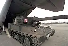 An AGS in woodland camouflage pattern rolls out of the rear cargo ramp of a C-130. Gun is forward. There is no armament mounted on the commander's hatch.