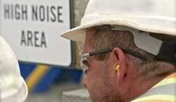A construction worker wearing hearing protection in front of a sign which reads